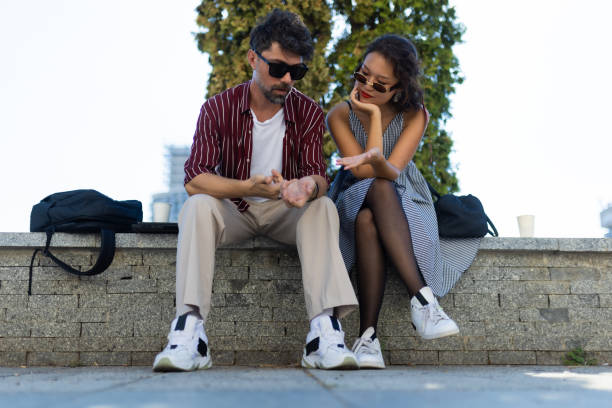 A man and woman sit on a wall outdoors, engaged in a focused conversation on a sunny day, dressed casually