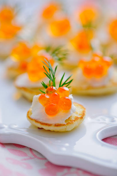 Close-up of mini blinis topped with sour cream, salmon roe, and fresh dill on a decorative white platter