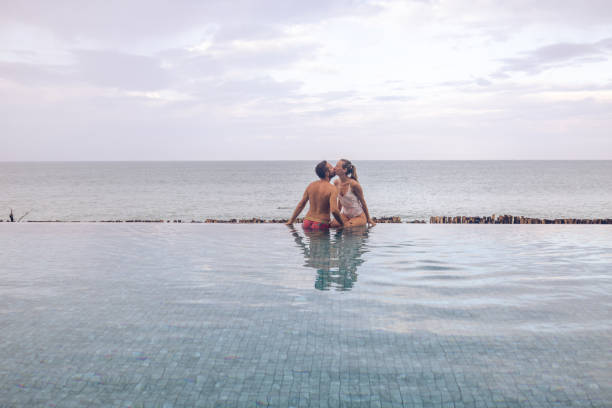 A couple sits on the edge of an infinity pool, kissing with the ocean horizon in view. The serene setting captures a moment of relaxation and romance by the sea.