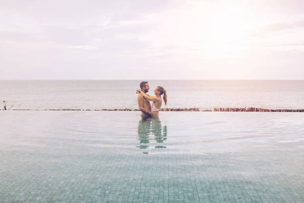 A cheerful couple spending quality time by the sparkling pool in warm sunlight, enjoying a tranquil and romantic setting adorned with relaxing and natural surroundings.