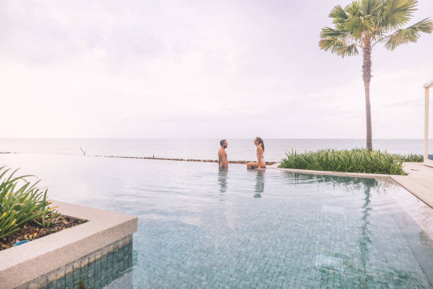 Romantic scene of a couple sitting by an infinity pool overlooking the ocean. Serene atmosphere with palm tree and calm waters, ideal for relaxation and leisure.