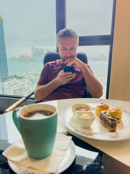 Man relaxing by window, browsing on phone with dessert and coffee.