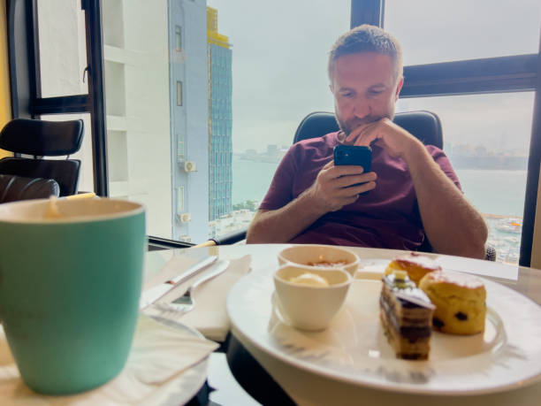 Man relaxing by window, browsing on phone with dessert and coffee.