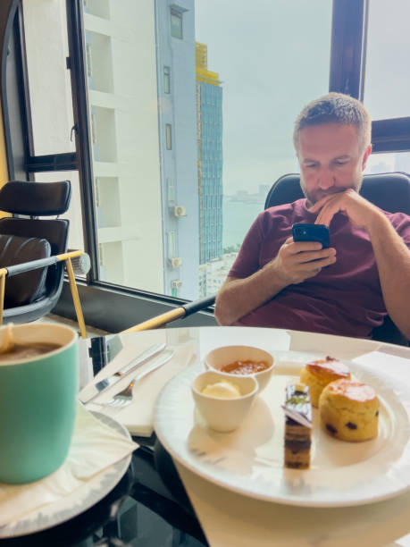 Man relaxing by window, browsing on phone with dessert and coffee.