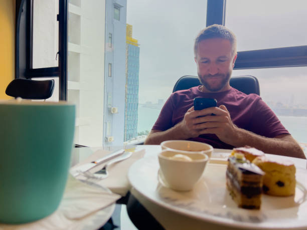 Man relaxing by window, browsing on phone with dessert and coffee.