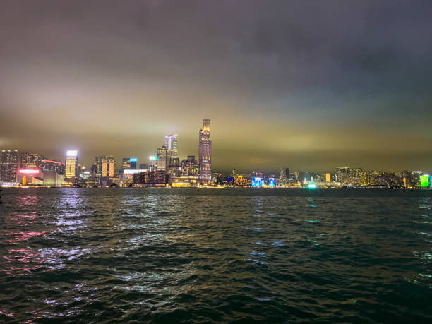 City skyline glowing at night seen from waterfront. Tall buildings reflect on rippling water surface. Urban landscape with vibrant city lights and peaceful night setting.