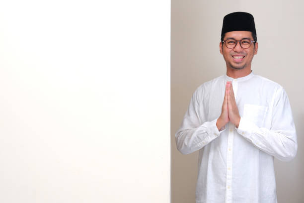 A muslim man standing beside blank board doing hand praying gesture with happy face