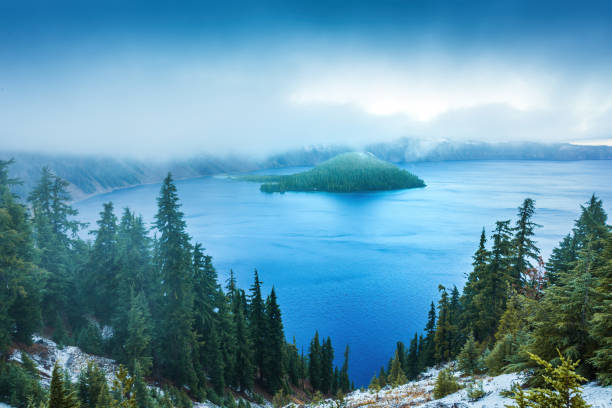 Misty morning shrouds Crater Lake National Park in Oregon, highlighting the serene, deep blue waters, evergreen forest, and iconic island, creating a tranquil and mystical atmosphere.