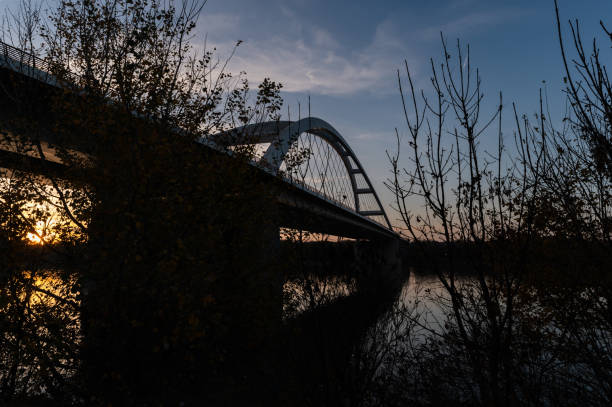 A captivating silhouette of the Pentele Bridge captured at dusk, with the sunset reflecting on the calm waters of the river. The bare trees in the foreground enhance the serene atmosphere.