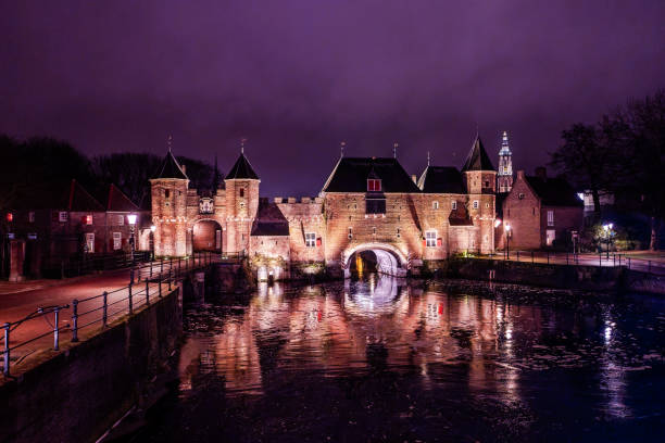 Amersfoort, The Netherland. December 8th, 2024: A stunning nighttime view of the Koppelpoort in Amersfoort, The Netherlands. The medieval city gate is beautifully illuminated, with its reflection shimmering on the calm water of the canal. The iconic Onze Lieve Vrouwetoren can be seen in the background, adding depth to this picturesque scene.