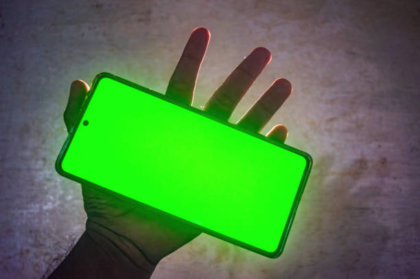 A hand holding a smartphone with a bright green screen, creating a glowing effect. The vibrant green display contrasts with the dim background, making the image visually striking