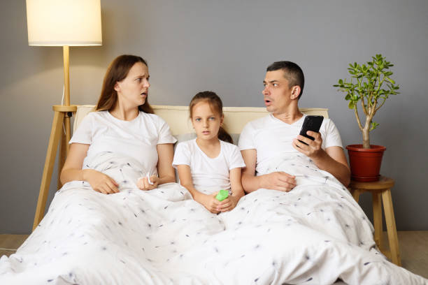 istock A surprised mother and father sit on a bed covered with blankets, staring at a phone while their curious daughter sits between them, all in a modern living space 2188993664