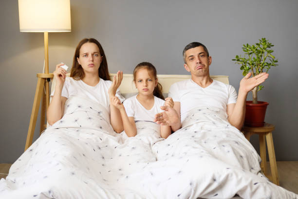 istock A family of three sits together on a bed, looking puzzled while facing a television. The parents display expressions of uncertainty, with the child appearing equally confused 2188993694
