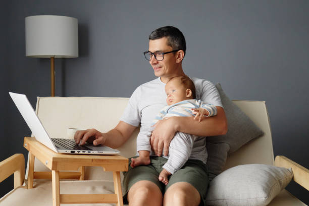 istock A father is sitting on a couch, working on his laptop while gently holding his sleeping baby. The setting is a cozy living room featuring neutral colors and soft lighting 2188996029