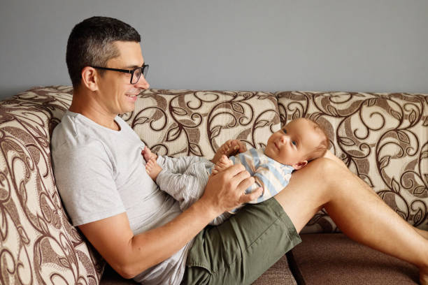 istock A father enjoys quality time with his smiling baby, engaging in playful interactions while seated comfortably on a patterned couch in a cheerful living room 2188996150