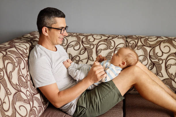 A father sits on a sofa with a baby resting on his legs, both smiling and interacting happily. The relaxed atmosphere captures a tender bonding moment in their cozy living room