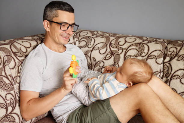 istock A father joyfully interacts with his baby while sitting on a comfortable couch. The father holds a colorful toy, capturing the baby's attention during playtime 2188996152