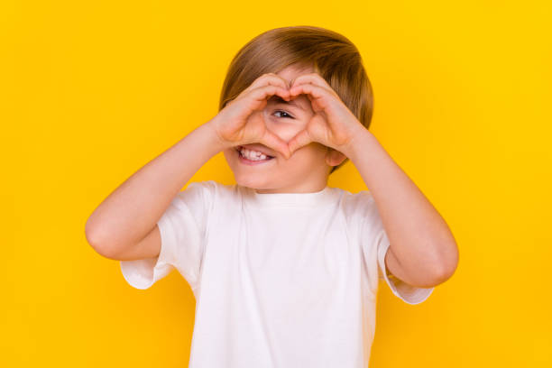 Portrait of handsome funny cheerful pre-teen boy looking through heart symbol isolated over bright yellow color background Portrait of handsome funny cheerful pre-teen boy looking through heart symbol isolated over bright yellow color background. cute boy show love symbol hand stock pictures, royalty-free photos & images