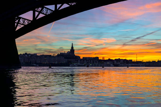 Skyline of the city Nijmegen on the riverbank of the Waal seen from under the Waalbrug during sunset. Various ships are moored at the quay of the river during this beautiful cold winter sunset during the blue hour at the end of the day.