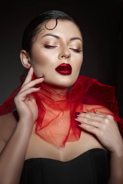 Model poses gracefully with eyes closed, highlighting striking red lipstick and makeup. sheer red scarf drapes around her neck, creating a dramatic and sophisticated look.