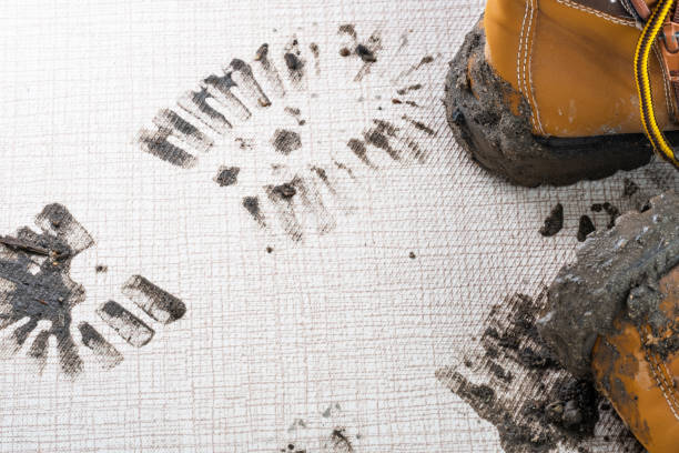 A close up of the wet muddy footprints on the carpet or floor in the living room. A pair of brown boots on the background. Concept of cleaning and dirty stains