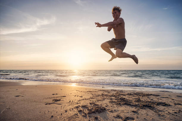 Shirtless, strong boy is training karate on a sandy beach. He is concentrating intently, moving with precision and strength from one martial arts pose to another as he practices his kata routine, demonstrating remarkable skill. The boy is captured mi-air, performing the jump of Enpi kata..Shot with Canon R5