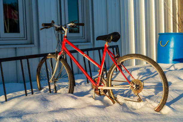 A vibrant red bicycle is parked in deep snow near a building, casting long shadows on a sunny winter day. The scene captures the contrast between the bright bike and white snow.