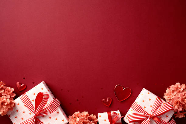 Valentines Day concept with gift boxes wrapped in heart-patterned paper and red ribbons, surrounded by pink flowers and heart-shaped decorations on a red background. Perfect for romantic celebrations