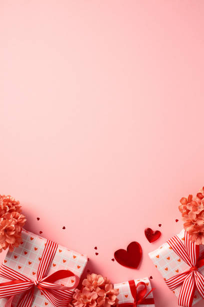 Valentine's Day flat lay with gift boxes wrapped in heart-patterned paper and red ribbons, surrounded by red flowers and heart-shaped decorations on a pink background