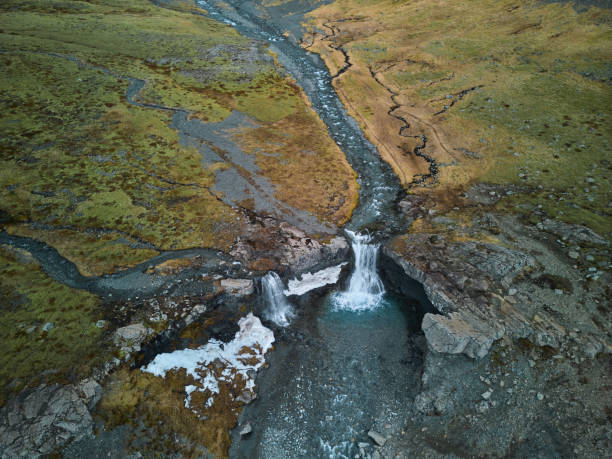 Aerial view of two waterfalls flowing down a mountain river, creating a small lake in a rocky landscape covered with green and yellow grass