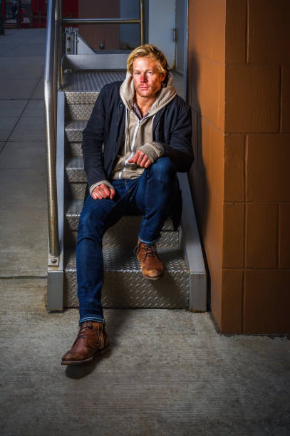 A person sits on metal steps, wearing jeans and brown boots, with a thoughtful expression on his face. The scene takes place outdoors next to a brown brick wall, with a moody urban backdrop.