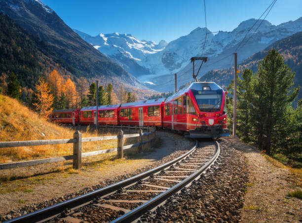 roter moderner elektrozug, der auf einer eisenbahn fährt und sich durch ein atemberaubendes hochgebirge mit schneebedeckten gipfeln und leuchtenden herbstbäumen in der schweiz schlängelt. bernina express im herbst. eisenbahntourismus - schweiz stock-fotos und bilder