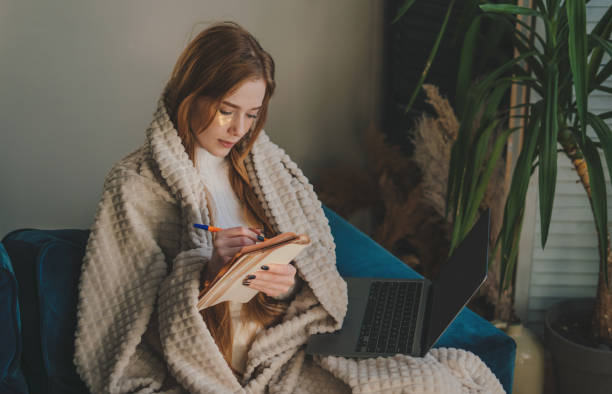 istock A cozy woman is comfortably writing while wrapped in a blanket, using her laptop 2189085115