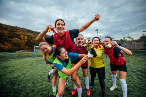 A team of female soccer players are celebrating a win of their team, jumping on each other and holding up a trophy and medals around their necks.
