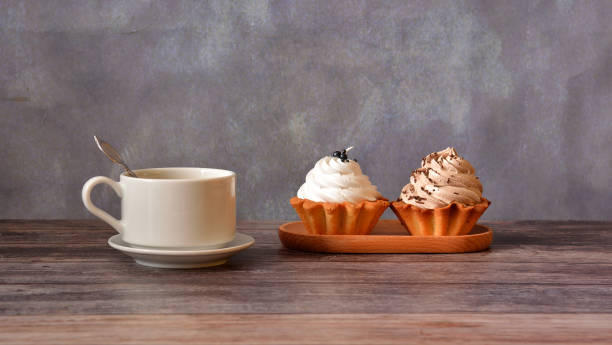 A cup of black coffee on a saucer with a spoon and an oval wooden plate with two shortbread cakes and a basket of protein cream on a wooden table.
