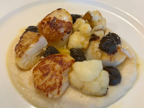 Full frame image of white plate containing restaurant appetiser of pan-fried scallops, cauliflower and capers on a bed of celeriac purée, elevated view, focus on foreground stock photo