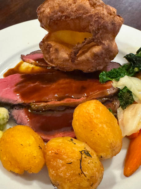 istock Full frame image of white plate containing Sunday roast dinner meal, Yorkshire pudding, carrots, roasted potatoes, cabbage, creamed leeks, slices of beef, covered in gravy, wood grain table background, elevated view, focus on foreground 2189124228