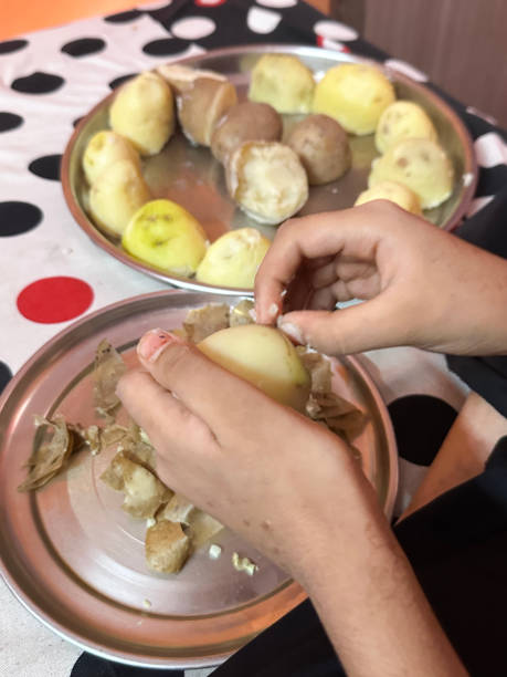 Stock photo showing close-up, elevated view of cooked boiled potatoes being peeled by hand by an unrecognisable Indian girl who is sat cross-legged on the floor wearing casual clothing.