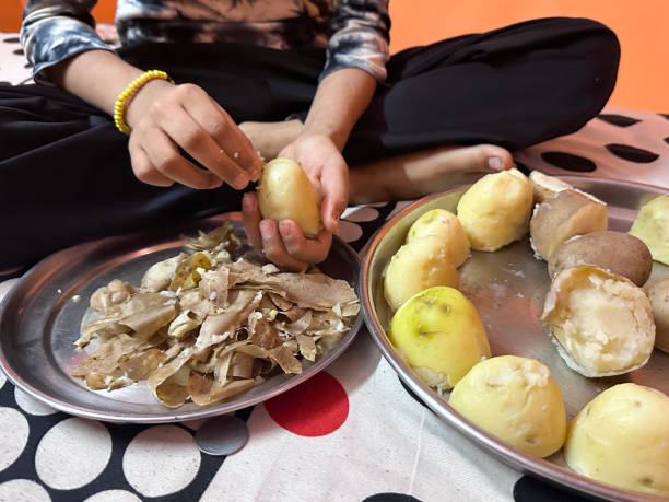 istock Close-up image of unrecognisable girl sat cross-legged on floor, preparing dinner, peeling cooked boiled potatoes by hand, placing skins onto stainless steel thali plate, focus on foreground 2189124657
