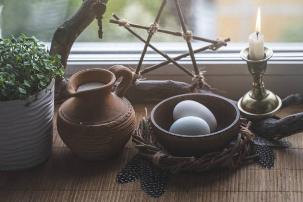 Magic home set. Atmospheric altar on windowsill with eggs, pentacle, the first greens in the pot, candle, birds feathers and clay jug with milk, selected focus. Ostara, Imbolc, Easter, March, spring.