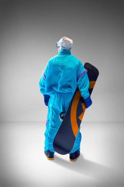 istock Rear view of athlete dressed in blue retro-inspired suit posing with snowboard against grey studio background. Ideal for extreme sports themes. 2189147635