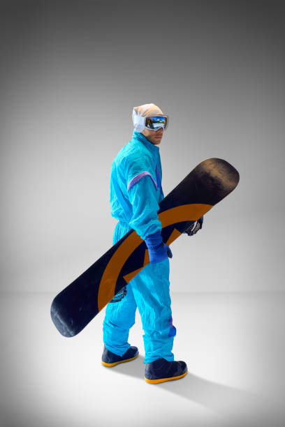 istock Side view of athlete dressed in blue retro-inspired suit posing with snowboard against grey studio background. Ideal for extreme sports themes. 2189147643
