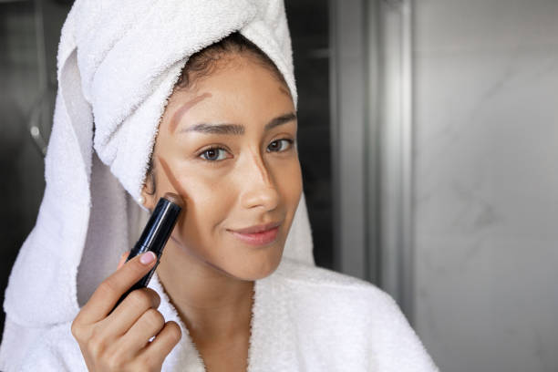 Smiling young woman applying contouring makeup stick on her face in bathroom