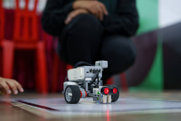 A close-up of a robot with wheels and sensors placed on a white surface. In the background, a person is kneeling down and focusing on the robot. The image captures a moment during a robotics session, with a blurred classroom setting in the background. The robot is designed for educational purposes, possibly for teaching kids or students about programming and engineering.