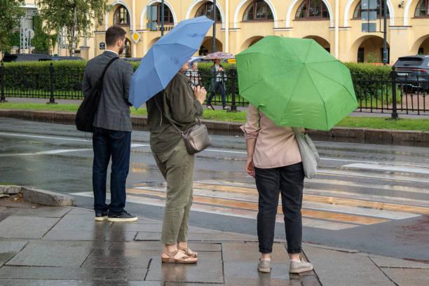 Saint-Petersburg, Russia - 2 August 2024. Three people are standing on a wet street, each holding a different colored umbrella. The scene is dreary and rainy, with the umbrellas providing some protection from the downpour