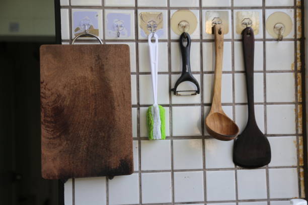 A wooden cutting board sits on a wall next to a rack of kitchen utensils. The utensils include a wooden spoon, a wooden spatula, and a wooden peeler. The cutting board is slightly burnt