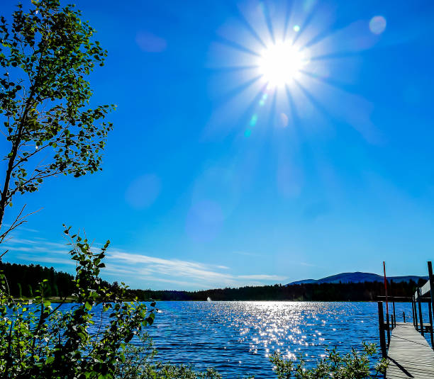 A beautiful blue sky with a sun shining brightly over a lake. The water is calm and the trees are lush and green