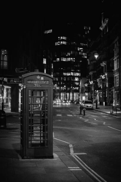 London, England, UK – January 15, 2024: A vintage red phone booth stands alone on a quiet London street, surrounded by modern buildings and illuminated by soft streetlights. This black-and-white photo captures the timeless charm of the city.