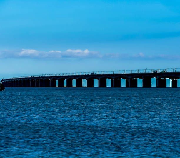 A long bridge spans a body of water. The sky is clear and blue. The bridge is lit up at night