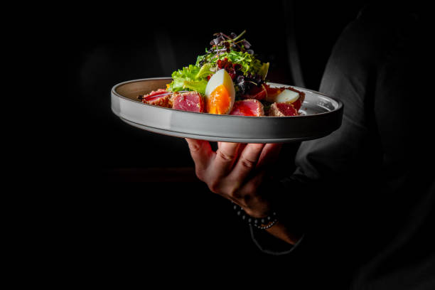 istock A hand elegantly presents a gourmet dish featuring fresh greens, sliced meats, and a perfectly cooked egg, set against a dramatic dark background. Ideal for food photography. 2189235812